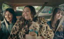 Three women sit in the backseat of a car, laughing. The one in the middle has a small dog on her lap.