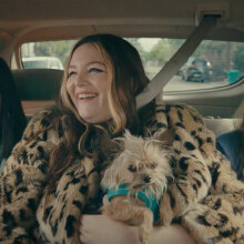 Three women sit in the backseat of a car, laughing. The one in the middle has a small dog on her lap.