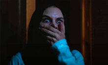 A woman stands in a dark room with her hand pressed to her mouth, looking terrified.