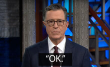 A man in a suit stands on a talk show stage staring at the camera with a neutral expression. The caption below reads: "Okay."