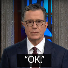 A man in a suit stands on a talk show stage staring at the camera with a neutral expression. The caption below reads: "Okay."