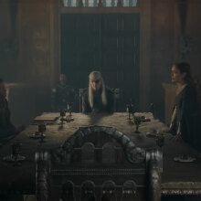 A group of people in medieval clothes sit around a large table in a gloomy room.