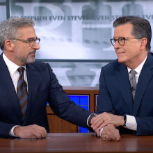 Two men in suits sit behind a desk, looking at each other with affection.