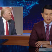 A man in a suit sits behind a talk show desk. In the top-right corner is an image of another man.