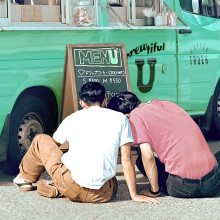 Two men sitting next to each other in front of a coffee truck. Their backs are to the viewer.