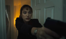 A close-up of a woman walking into a room with a gun raised in both hands.