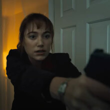 A close-up of a woman walking into a room with a gun raised in both hands.
