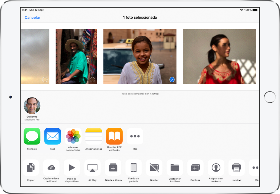 Pantalla Compartir con fotos en la parte superior. Hay una foto seleccionada, que se indica con una marca de verificación blanca en un círculo azul. La fila situada debajo de las fotos muestra amigos con los que puedes compartirlas mediante AirDrop. Debajo hay otras opciones para compartir, entre las que se incluyen, de izquierda a derecha, Mensajes, Mail, “Álbumes compartidos”, “Añadir a Notas”, “Guardar PDF en Libros” y Más. En la fila inferior se encuentran los botones Copiar, “Guardar en Archivos”, “Copiar enlace de iCloud”, “Pase de diapositivas”, AirPlay, “Añadir a álbum”, “Fondo de pantalla”, Ocultar, Duplicar, “Asignar a un contacto”, Imprimir y Más.