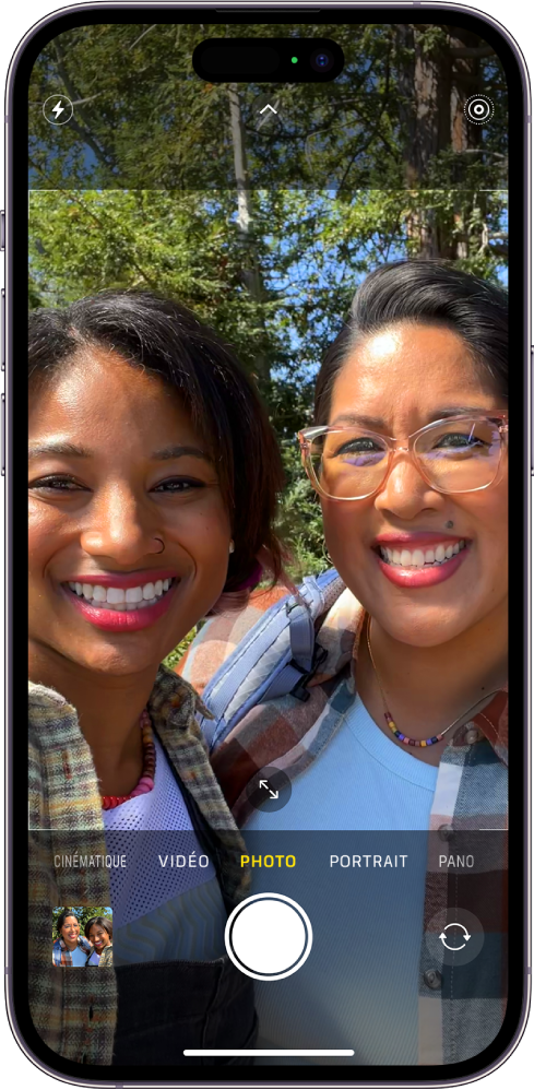 L’écran « Appareil photo » en mode Photo. En dessous des différents modes d’appareil photo, de gauche à droite, se trouvent le bouton « Visualiseur de photos et vidéos », le bouton « Prendre une photo » et le bouton « Sélecteur de caméra - face arrière ». Les boutons pour le flash, les commandes de l’appareil photo et le mode Live Photos figurent en haut de l’écran. Dans le cadre de l’appareil photo, les sujets prennent un selfie.