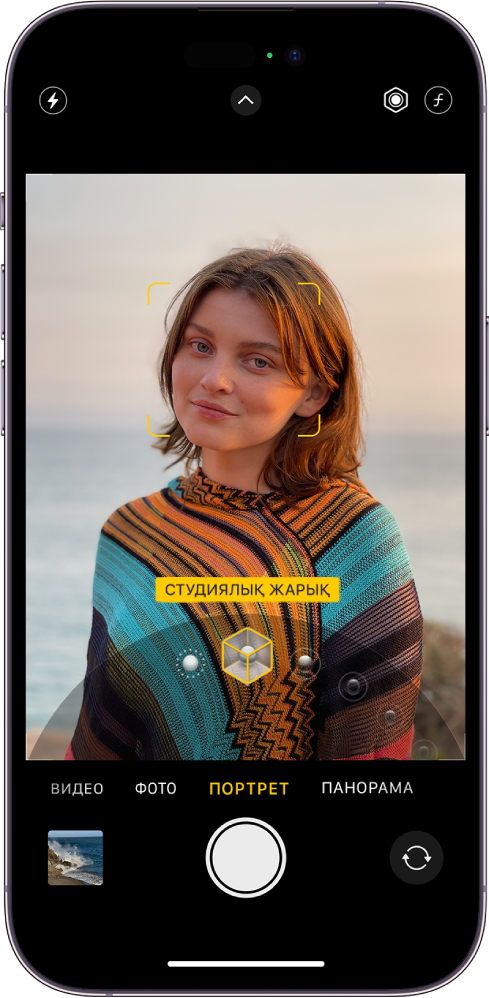 «Портрет» режиміндегі «Камера» экраны; көрініс тапқышта нысан анық және өң бұлыңғыр. «Портреттік жарықтандыру» параметрін таңдау үшін диск жақтаудың төменгі жағында ашық және «Студиялық жарық» параметрі таңдалған. Экранның жоғарғы сол жағындағы — «Жарқыл» түймесі, жоғарғы ортада — «Камераны басқару элементтері» түймесі, ал жоғарғы оң жақтағылар — Портрет жарығының қарқындылығын реттеу және «Тереңдікті басқару» түймелері. Экранның төменгі жағындағылар солдан оңға қарай — «Фото және видео көру құралы» түймесі, «Суретке түсіру» түймесі және «Камера таңдау - артқы камера» түймесі.