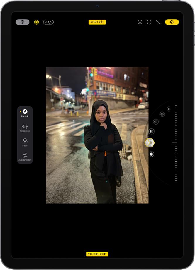 Der Bildschirm „Bearbeiten“ für ein Foto im Porträtmodus in der App „Fotos“. Das Foto wird in der Bildschirmmitte angezeigt. Rechts befinden sich die Optionen für den Porträtlichteffekt. Die Taste „Studiolicht“ ist ausgewählt und ein Schieberegler ermöglicht die Anpassung der Effektintensität. Auf der linken Seite des Bildschirms befinden sich die Tasten „Porträt“, „Anpassen“, „Filter“ und „Zuschneiden“. Die Taste „Porträt“ ist ausgewählt.