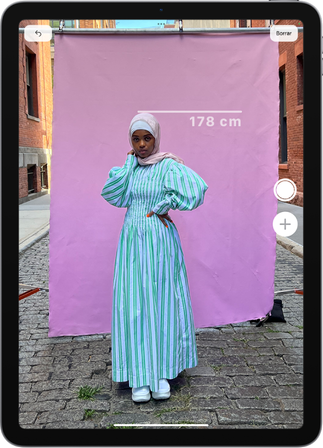 Medida de la altura de una persona en la app Medidas, con el valor mostrado en la parte superior de la cabeza de la persona. El botón “Hacer foto” está activo cerca de la esquina inferior derecha para hacer una foto de la medición. El indicador verde “Cámara en uso” aparece en la parte superior.