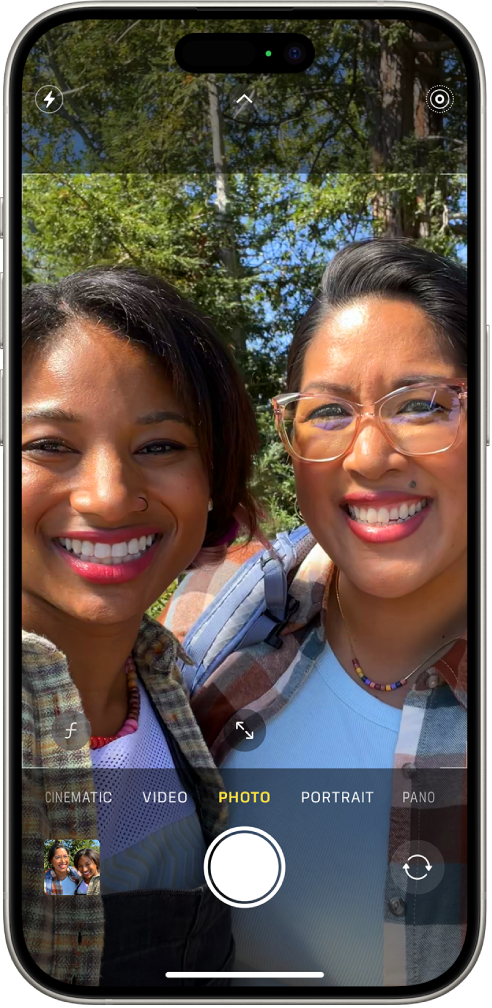 Pantalla de l’app Càmera en mode de foto. A sota dels modes de la càmera, d’esquerra a dreta, hi ha el botó visor de fotos i vídeos, el botó per fer una foto i el botó selector de la càmera posterior. A la part superior de la pantalla hi ha els botons del flaix,dels controls de la càmera i de Live Photos. Dins el marc de la càmera, els subjectes estan fent una selfie.