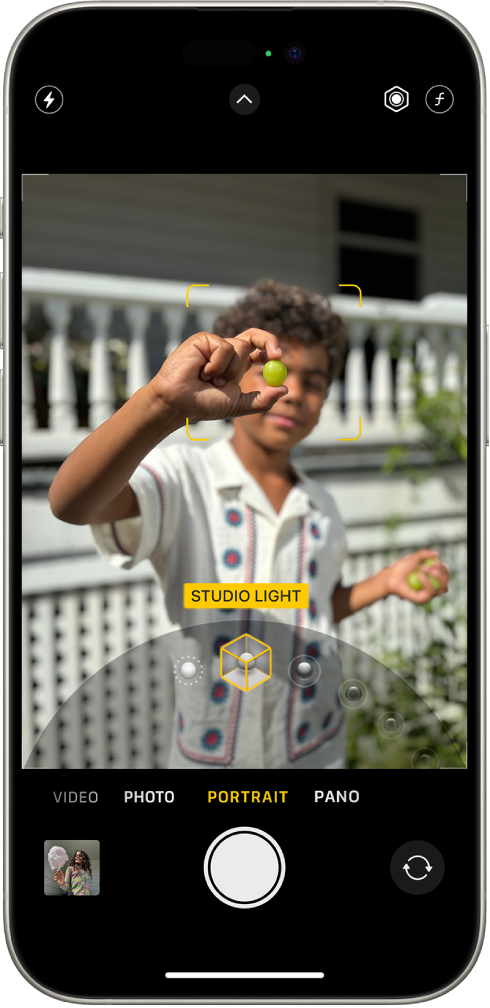 Lietotnes Camera ekrāns režīmā Portrait; skatu meklētājā objekts ir fokusā, un fons ir aizmiglots. Kadra apakšdaļā atvērta skala, kas ļauj izvēlēties režīma Portrait Lighting efektus, un ir atlasīts efekts Studio Light. Ekrāna augšējā kreisajā stūrī ir poga Flash, centrā ir poga Camera Controls, un augšējā labajā stūrī ir pogas, kas ļauj pielāgot režīma Portrait Lighting intensitāti un Depth Control. Ekrāna apakšā no kreisās puses uz labo ir pogas Photo and Video Viewer, Take Picture un Camera Chooser Back-Facing.