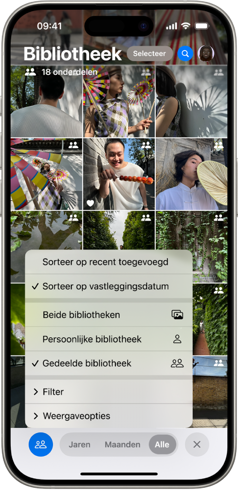 Een gedeelde iCloud-fotobibliotheek in de Foto's-app. De knop 'Gedeelde bibliotheek' linksonderin is geselecteerd en 'Gedeelde bibliotheek' is geselecteerd in een lijst met bibliotheekopties. Bij alle foto's die in de bibliotheek worden weergegeven, staat in de rechterbovenhoek de badge voor 'Gedeelde bibliotheek'.