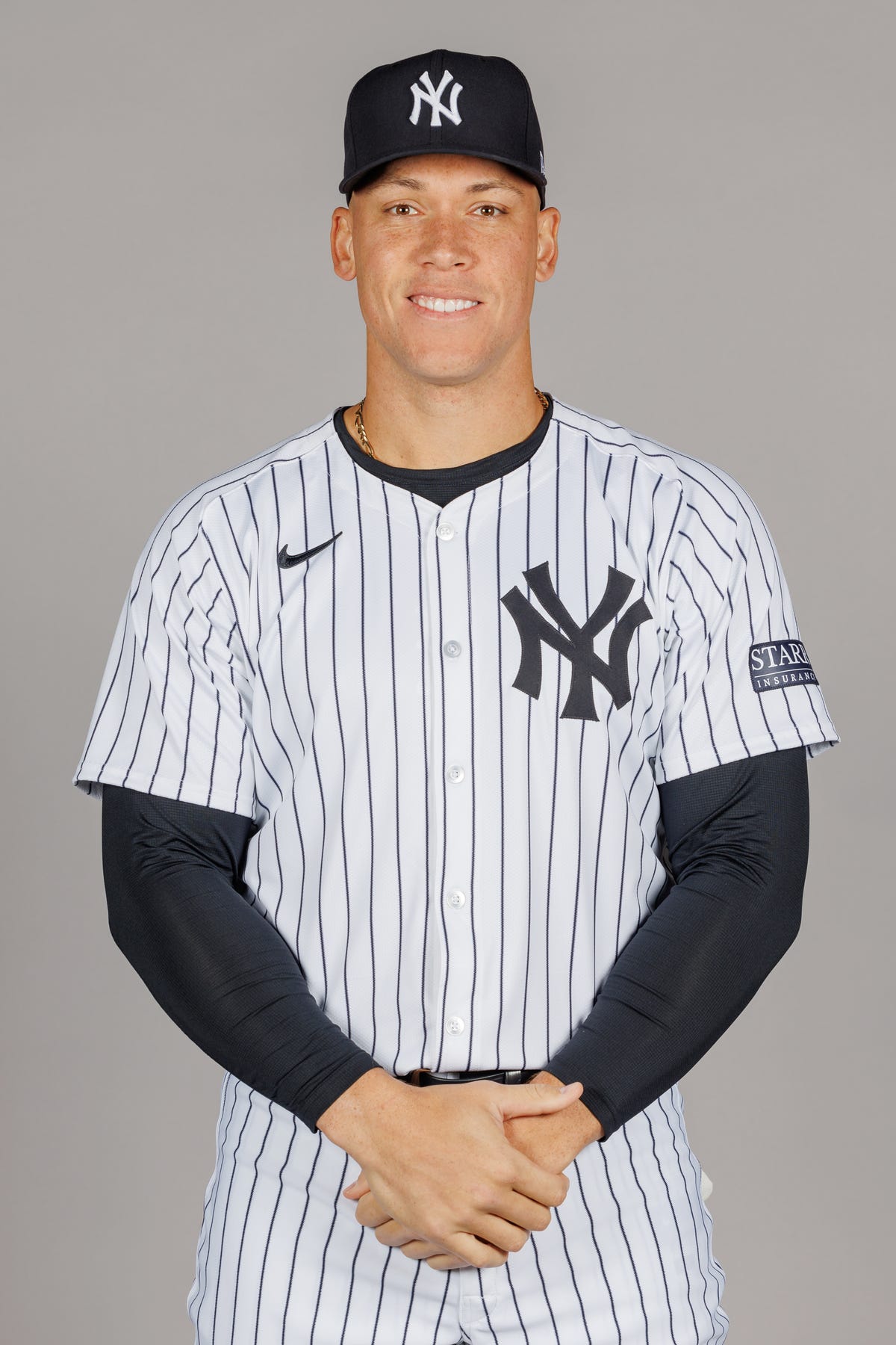 aaron judge smiles at the camera, he wears a white pinstripe new york yankees jersey and navy yankees baseball hat with a dark undershirt