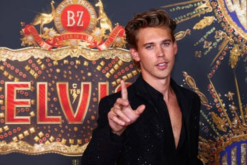 austin butler wearing a black shirt, holding a finger in the air, and standing in front of a logo with the word elvis on it