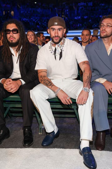 bad bunny looks at the camera while sitting down next to people, he wears a white t shirt and jeans with jewelry and a backward baseball cap