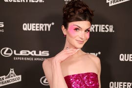 dylan mulvaney wearing a pink dress and blush makeup, putting her right hand under her chin, smiling directly at the camera