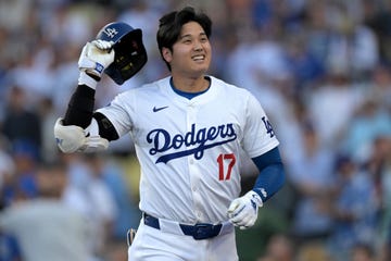 shohei ohtani smiles as he pulls a hat off his head, he wears a white los angeles dodgers baseball uniform including gloves and an elbow pad