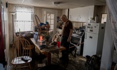 A man in a T-shirt stands in a kitchen with a chair on a table and mud on the floor