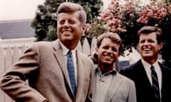 A color-tinted photo of three smiling white men wearing casual suits and standing in a row, looking in the same direction, in front of a pink rosebush and a white picket fence.