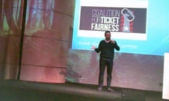 A man stands on stage in front of a banner saying 'Coalition for Ticket Fairness' in a still from hidden camera footage