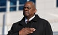 Black man without hat but wearing black trench coat over shirt and tie stands with hand over heart outside.