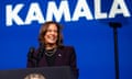 A woman stands at a podium and smiles with her name "Kamala" written on the screen behind her