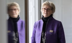 An older white woman wearing glasses and a purple jacket stands by a window that reflects her image