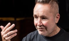 Violinist Nigel Kennedy, in a T-shirt and with a shaved-side haircut long on top, looks down slightly smiling gesturing with one hand