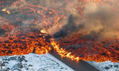 Huge amounts of lava rolls from snow-covered ground across a road and onto the snow-covered ground on the other side.