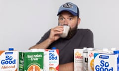 A bearded man wearing a cap drinking a glass of soy milk with a bewildered expression, with a selection of alternative milks laid before him on a table.