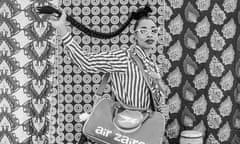 Black and white photo of a women with a long plaited ponytail, wearing a striped outfit and standing against a patterned background