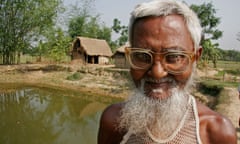 A Bangladeshi man wearing a pair of glasses smiles at the camera. Behind him is a fishpond and fields.