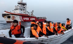 In the foreground, people wearing lifejackets are in a boat. In the background is the NGO's larger boat with the name 'Open Arms' on the side