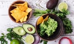 Top view of a bowl of guacamole, with avocado halves and lime halves to the side, with a bowl of corn tortilla chips nearby, on w white wooden surface.