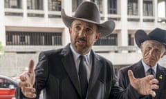 A middle-aged white man wearing a distinctive dark gray blazer and cowboy hat, whose tips turn up at the sides, speaks and gestures on a sidewalk. Has a beard.