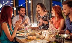 Three young men and two young women around a table with food and a lit birthday cake on it, smiling and clapping