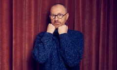 Mark Gatiss, standing in front of a red stage curtain and wearing a blue jumper, holds his fists up to his chin.