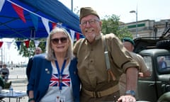 Suzy and Barry Shimell pose in front of military vehicles with Barry wearing an old-fashioned military uniform and Suzy wearing a Union Jack t-shirt