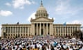 Long, white-stone building with taller columns in front and cupola beyond, with hundreds of people in white T-shirts gathered in front.