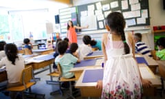 Little girl with light brown skin and long black hair in pink T-shirt under white floral dress, seen from behind in a grade school classroom, stands up at her desk in the back of a room with other students sitting at their desks.