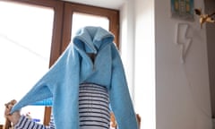 Child (10-11) standing in front of a large window in a bedroom and pulling a blue knitted sweater over her head to get dressed as part of the morning routine.<br>Stock image of a child putting on a jumper