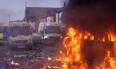 A vehicle ablaze on a litter-strewn street as police vans are parked nearby