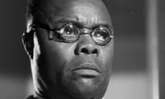 A close-up of a wide-faced black man in small round glasses reacting severely to something off-camera