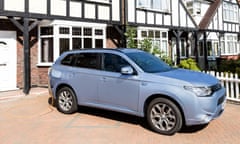 a silver-blue Mitsubishi Outlander PHEV, a large SUV, is seen side-on parked on a tiled driveway in front of a house with a mock-Tudor black and white frontage, white front door and windowframes, and white picket fence. A yellow cable is seen emerging from the charging port which is behind a flap in the car's rear wing, and this trails behind the car to the power source.