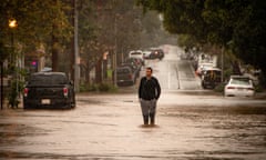 A middle-aged man with dark hair, wearing a sweater, pants, and knee-high boots, stands in the middle of an urban street with water up to his shins.