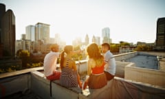 Group of friends on roof toasting at sunset<br>Group of friends sitting on roof ledge looking out at cityscape toasting wine glasses at sunset