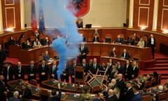 A man holds up a flare emitting bright blue smoke in a parliamentary chamber with seating arranged in a circle